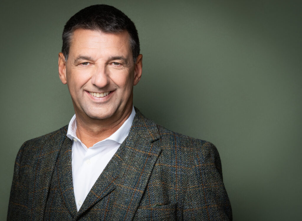 Peter Walleghem lachend in tweed jasje en open hemd tegen een groene achtergrond